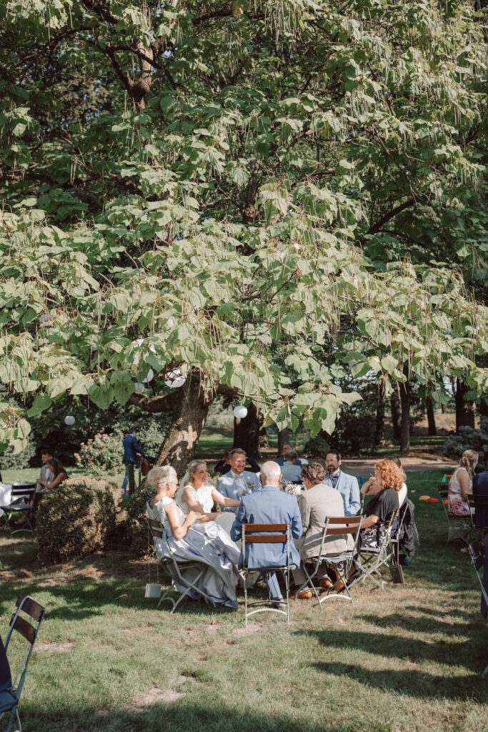 Hochzeitsgäste sitzen während der Feier unter einem großen Baum im Garten von Gut Wahnbek in Oldenburg – natürliche Hochzeitsfotografie von einer Hochzeitsfotografin aus Oldenburg.