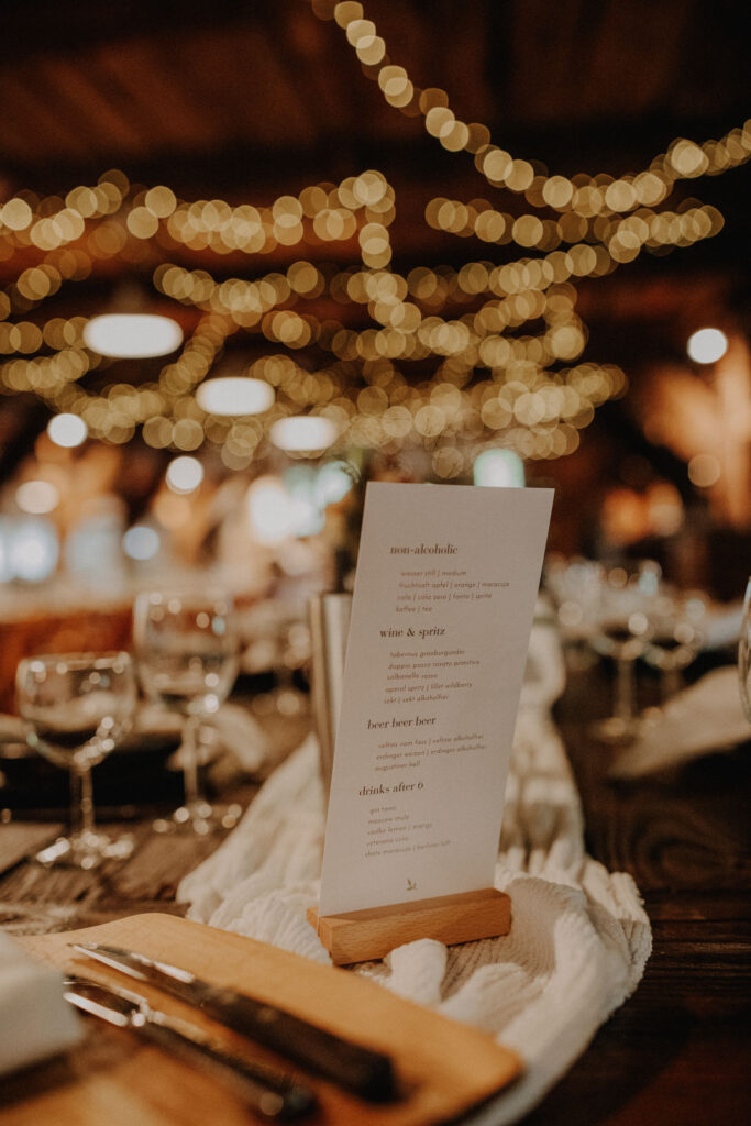 Stilvoll gedeckte Hochzeitstafel mit Menuekarte und stimmungsvoller Lichterdekoration in der Scheune des Hof Schweers. Authentische Hochzeitsfotografie und elegante Hochzeitsfotos vom Hochzeitsfotograf Oldenburg.