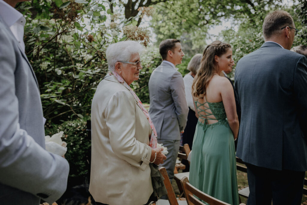 Hochzeitsgaeste verfolgen die freie Trauung im Garten des Hof Schweers. Authentische Hochzeitsfotografie und emotionale Hochzeitsmomente vom Fotograf Hochzeit Oldenburg.