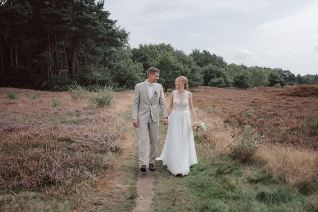 Braut und Braeutigam gehen Hand in Hand waehrend ihres Brautpaarshootings auf Gut Moorbek durch die Heidelandschaft. Natuerliche Hochzeitsfotografie und emotionale Hochzeitsfotos vom Hochzeitsfotograf Oldenburg.